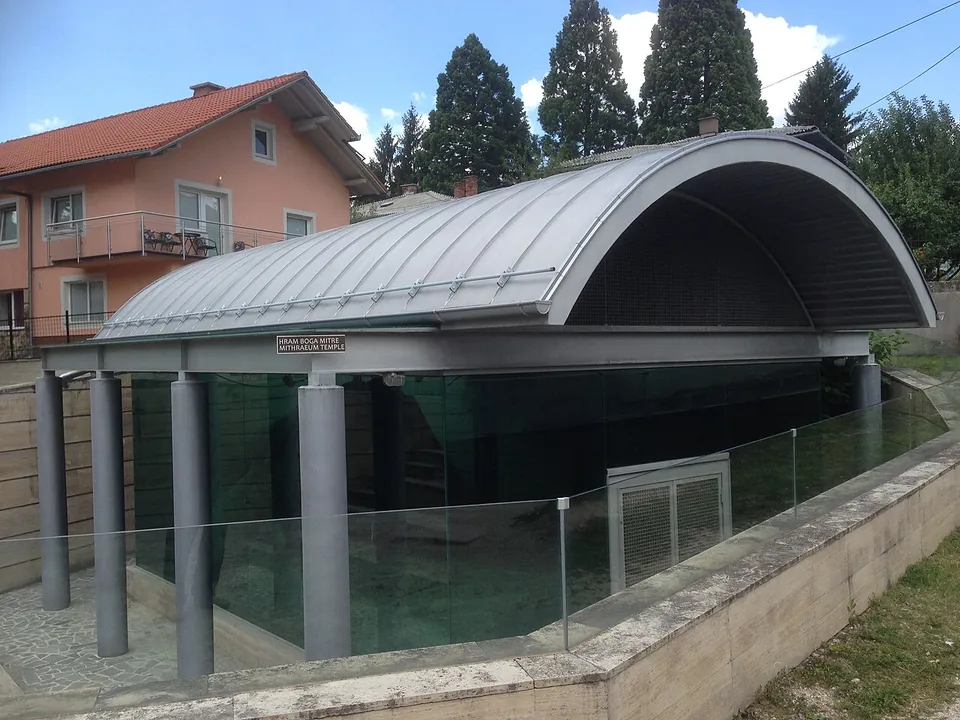 A small modern pavilion with a curved metal roof on grey columns and glass walls, standing below street level among houses and conifers.