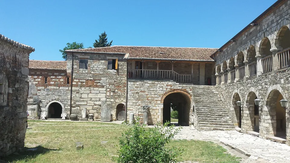 The courtyard of a stone monastery building with an arcaded upper gallery, a flight of steps and carved stones set around the lawn.