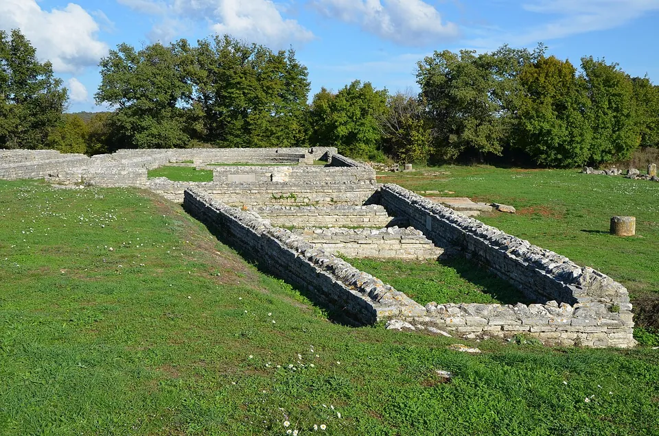 Low, neatly conserved stone walls outlining rectangular rooms in a mown meadow, with a line of oak trees behind under a blue sky with clouds.