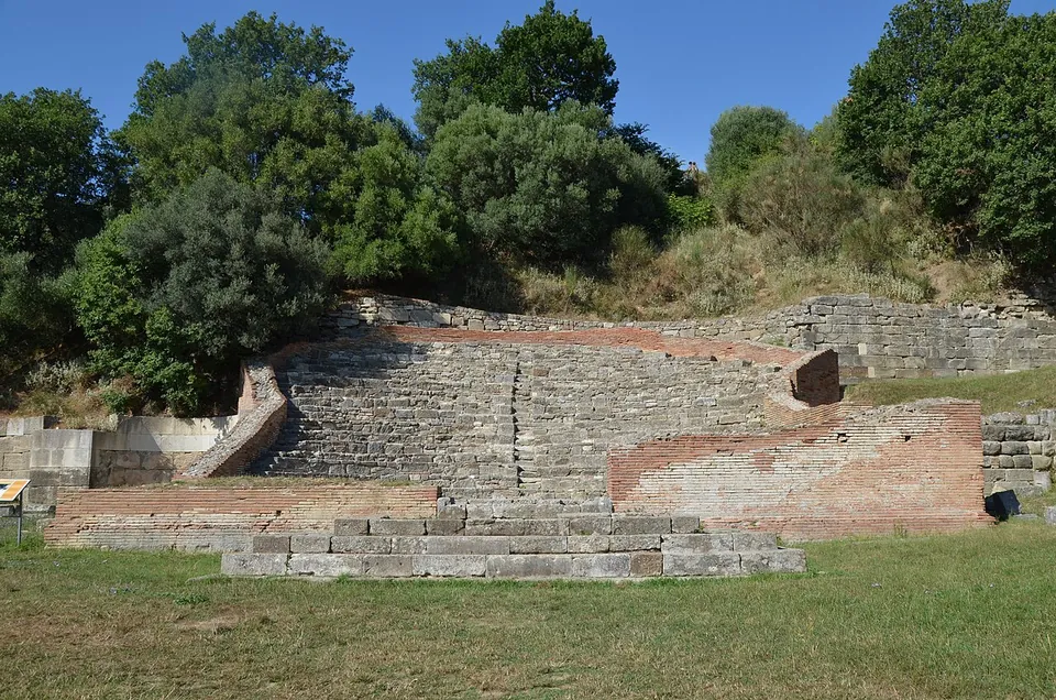 A small semicircular auditorium of stone seats rising behind a low stage platform, flanked by brick walls, set against a wooded slope.