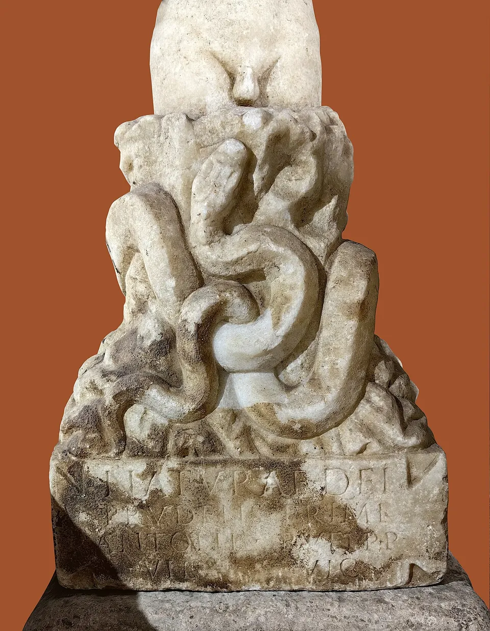 The lower half of a marble statue, a figure emerging from rough rock with a snake coiled around the rock, on a base with a worn inscription; the background is plain reddish brown.