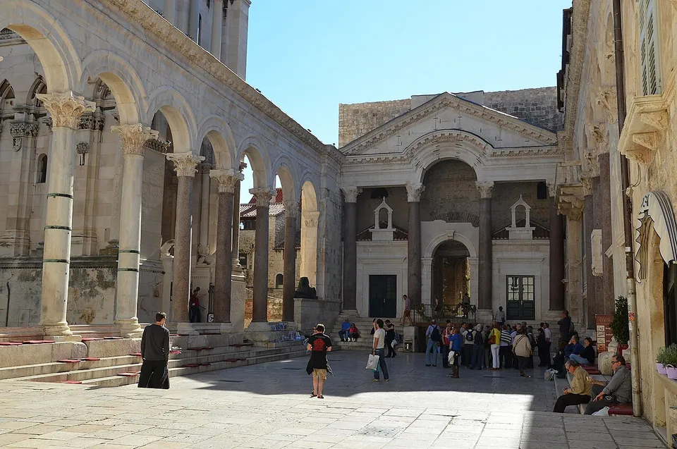 A paved Roman court lined on the left by an arcade of granite columns carrying round arches, closed at the far end by a pedimented front on four columns with an arched opening, with visitors standing in the sun.