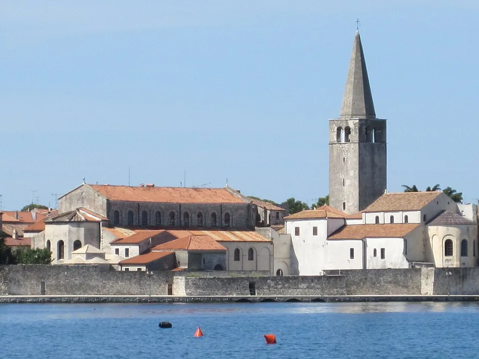 A cluster of pale stone church buildings with red-tiled roofs behind a low sea wall, dominated by a tall square bell tower with a pointed spire, seen across calm blue water.