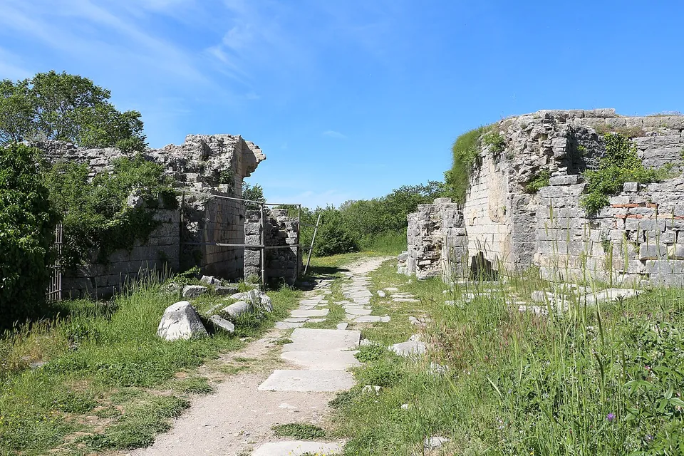 A stone-paved track running between the ruined masonry of two gate towers, overgrown with grass and shrubs, under a clear sky.