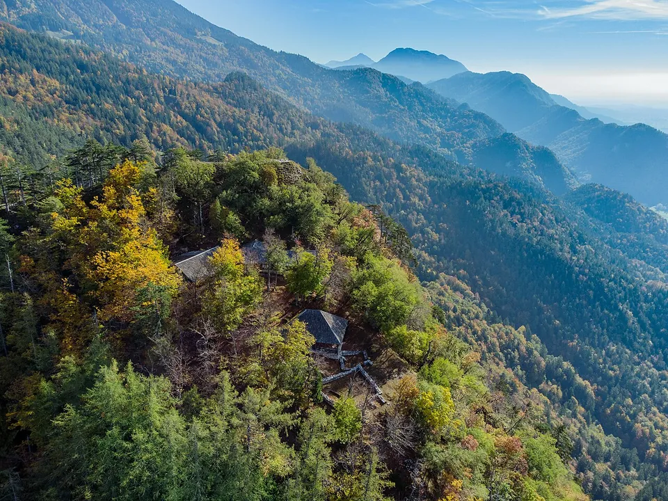 An aerial view of a wooded ridge top in autumn colour, with two shingle-roofed shelters standing among the trees on the summit and hazy mountain ranges receding behind.
