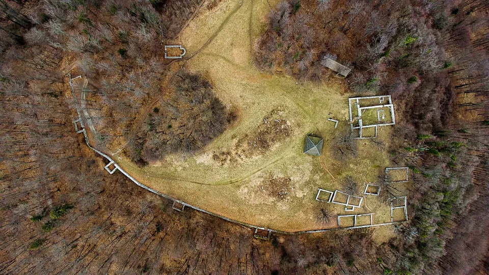 A hilltop photographed straight down from the air in winter, with a pale wall running round its edge and the rectangular outlines of buildings laid out on the bare grass inside, surrounded by leafless woodland.