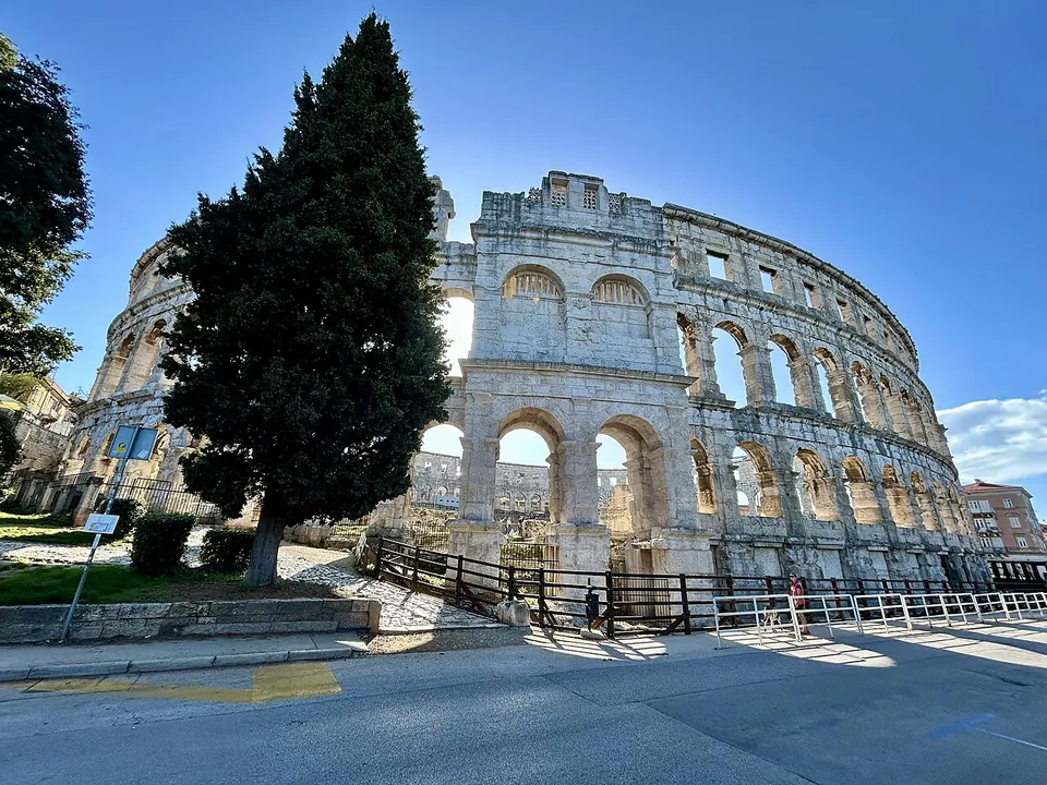 The pale limestone outer wall of the amphitheatre seen from the pavement, three storeys of arches and square windows rising against a blue sky, with a tall cypress in front.