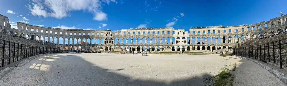 A wide panorama of the sandy oval floor inside the amphitheatre, ringed by a railing, with the arcaded outer wall encircling it under a blue sky.
