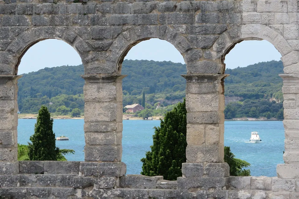 A row of three stone arches in the amphitheatre wall framing a view of blue sea, cypress trees and a green wooded shore.