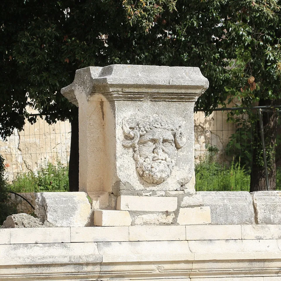 A weathered stone block carved with a bearded male head, standing on a low stone base in front of trees.