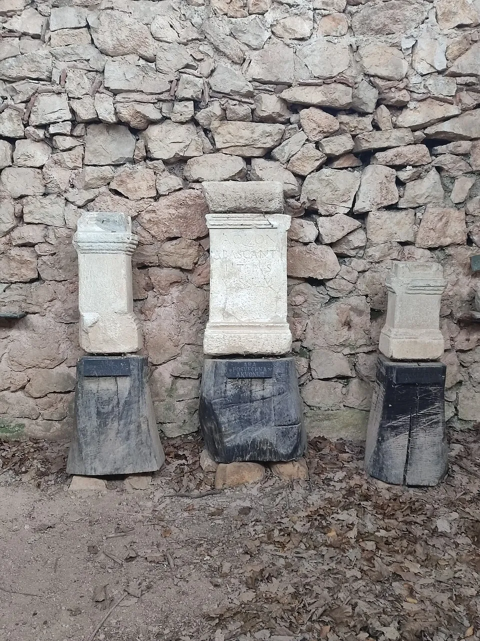 Three stone altars of different sizes standing side by side on dark plinths against a rough drystone wall, the middle one carrying a worn carved inscription.