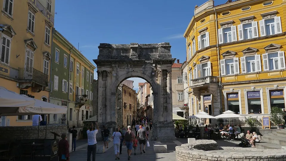 A single-arched Roman gateway of weathered stone standing at the end of a busy pedestrian street, between tall yellow and cream town houses with café awnings.