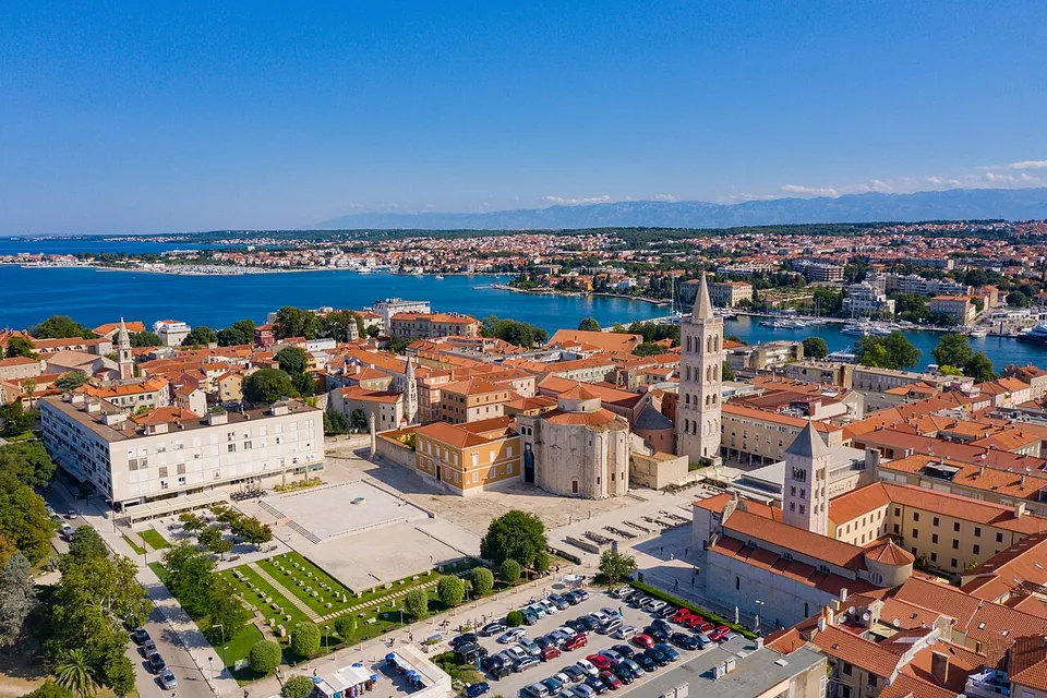 An aerial view of an old town square with a round domed church and a tall bell tower, the open area of the Roman forum beside it, a park and car park in the foreground and the sea behind.
