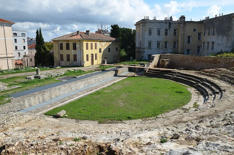 A semicircular Roman theatre on a hillside, its stone seating rows curving round a lawn, with a large yellow building and a pale villa above it and a low retaining wall along the stage side.