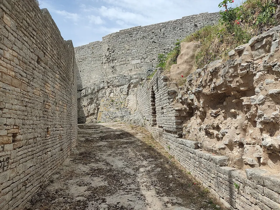 A narrow passage between a tall wall of neat stone blocks on the left and a rough rock face with masonry on the right, open to the sky.