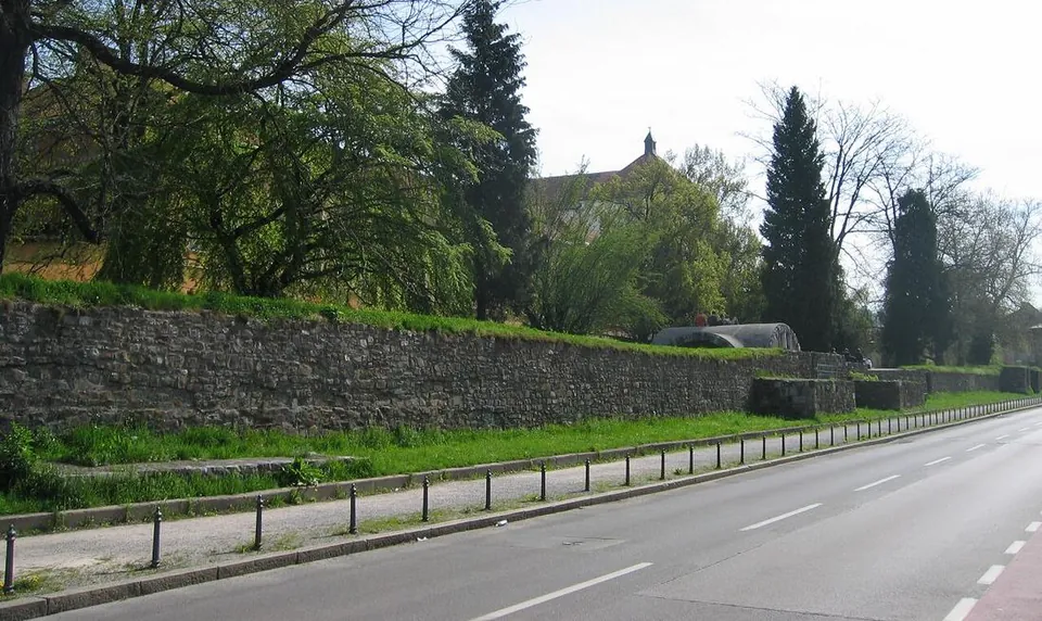 A long, low wall of grey rubble stone with turf along its top, running beside a pavement and a two-lane road, with trees and a church tower behind it.