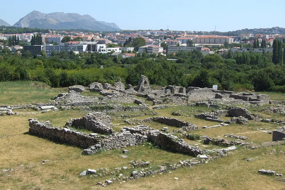 Low stone walls of excavated rooms spread across a dry field, with the buildings of the modern town and a mountain ridge behind.