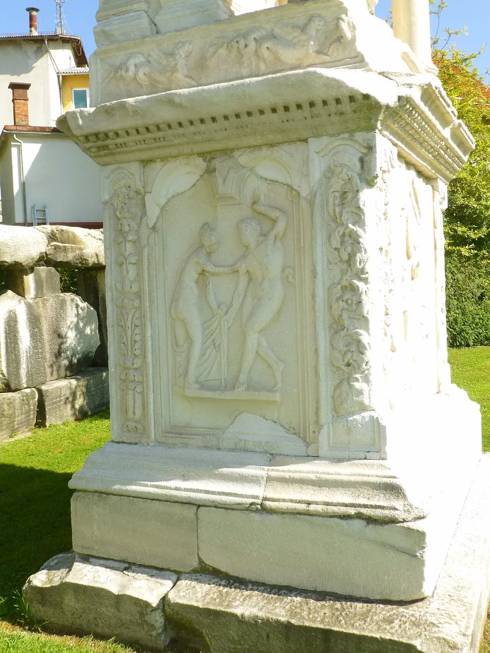 Close view of the corner of a white marble tomb base, a recessed panel carved with two figures in relief framed by carved vine scrolls, under a moulded cornice.