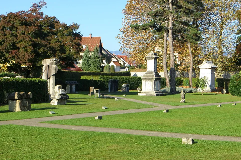 A mown park crossed by gravel paths and low hedges, with carved stone blocks, altar-like pillars and a tall white tomb monument scattered across it under autumn trees, houses behind.