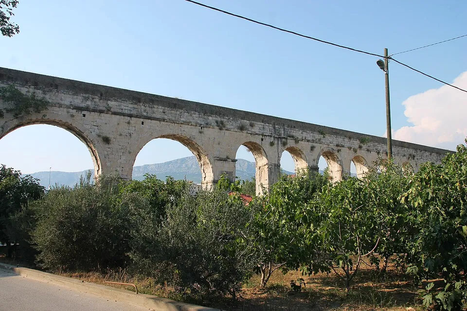 A line of stone arches carrying a flat-topped channel above olive and fig trees, beside a road with a lamp post, under a clear sky.