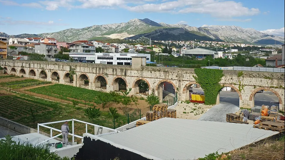 A long row of low stone arches running across a suburb between vegetable plots and a builder's yard, with modern houses and a bare limestone mountain behind.