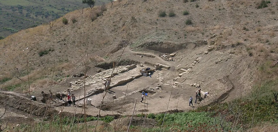 The curved hollow of an ancient theatre cut into a grassy hillside, with its lower stone rows and stage foundations exposed in an excavation, and a few people working.