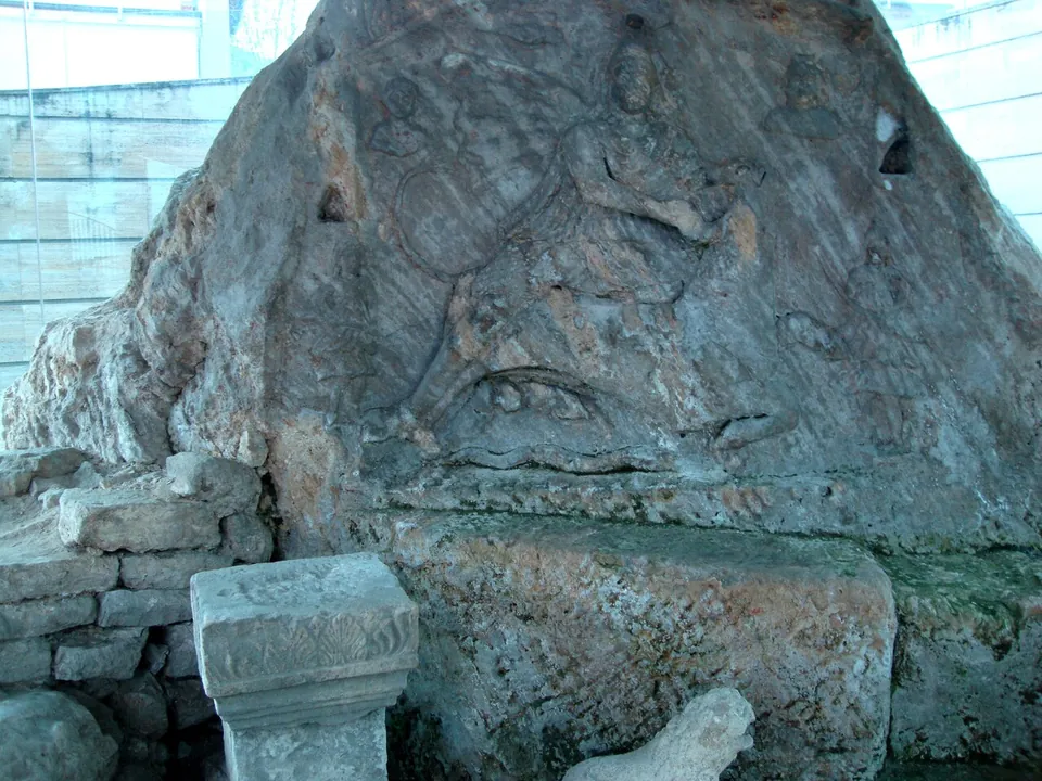 A large relief carved into a block of grey rock, worn but showing a figure kneeling on a bull, with a small carved stone altar and a sculpted animal fragment in front of it, seen through glass.