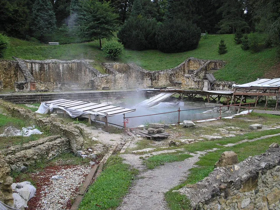 Roman stone walls several metres high along the foot of a green slope, with steam rising from a pool of water behind a railing and gravel paths among the ruins in the foreground.