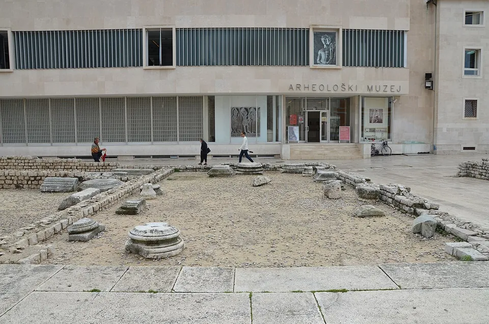 A sunken rectangle of rough ground with low stone wall lines and round column bases, edged by pale paving, in front of a modern stone museum building marked "Arheološki muzej".