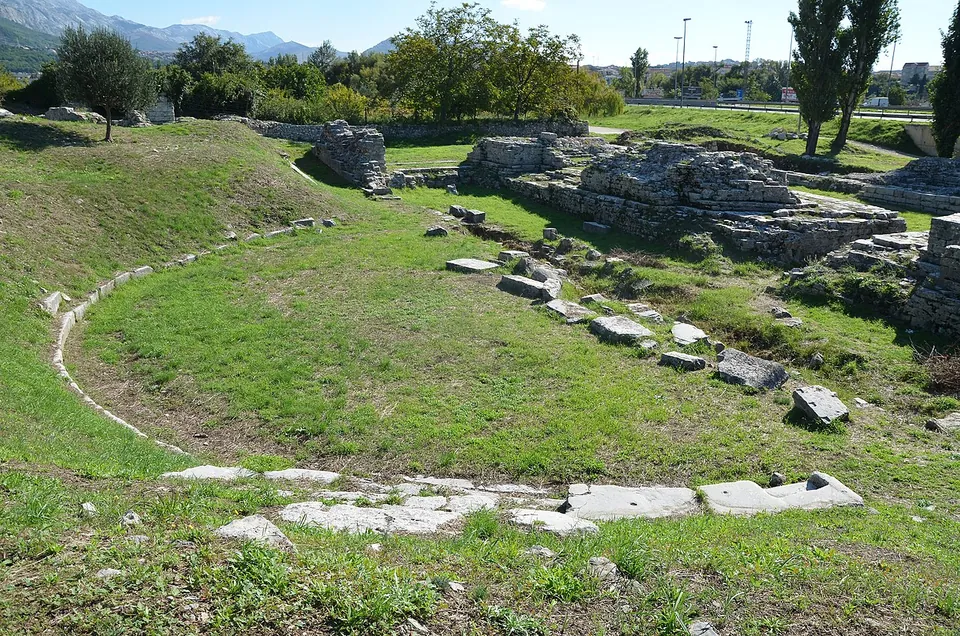 A grassy hollow shaped like a half circle, outlined by a curve of stones where the seating stood, with fragments of masonry and a road and trees beyond.