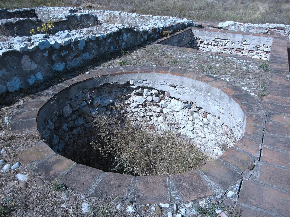 A deep circular shaft lined with rough stone, set into a floor edged with flat bricks, with low walls of stone and brick around it.
