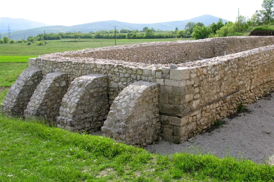 A rectangular block of conserved Roman walls of rough limestone, about head high, with a row of sloping buttresses along one side, standing in grass with fields and low hills behind.