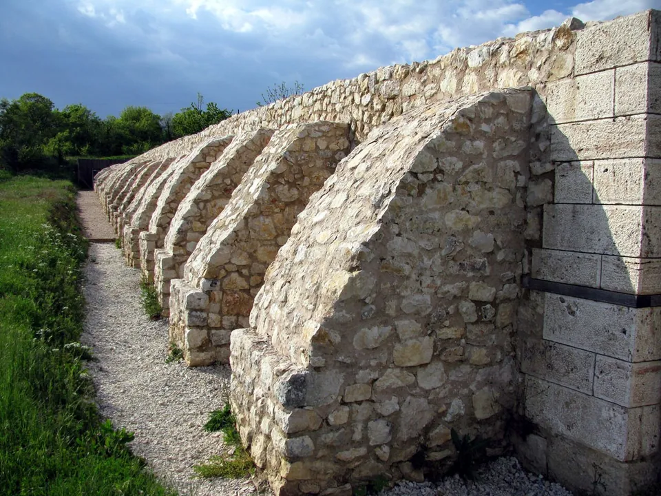 A long wall with a row of stone buttresses running away from the camera beside a gravel path, with a corner of squared blocks in the foreground and trees at the far end.