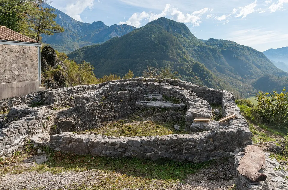 The rebuilt stone footings of a small building with a rounded end, knee to waist high, on a rocky hilltop, with steep forested mountains rising behind under a blue sky.