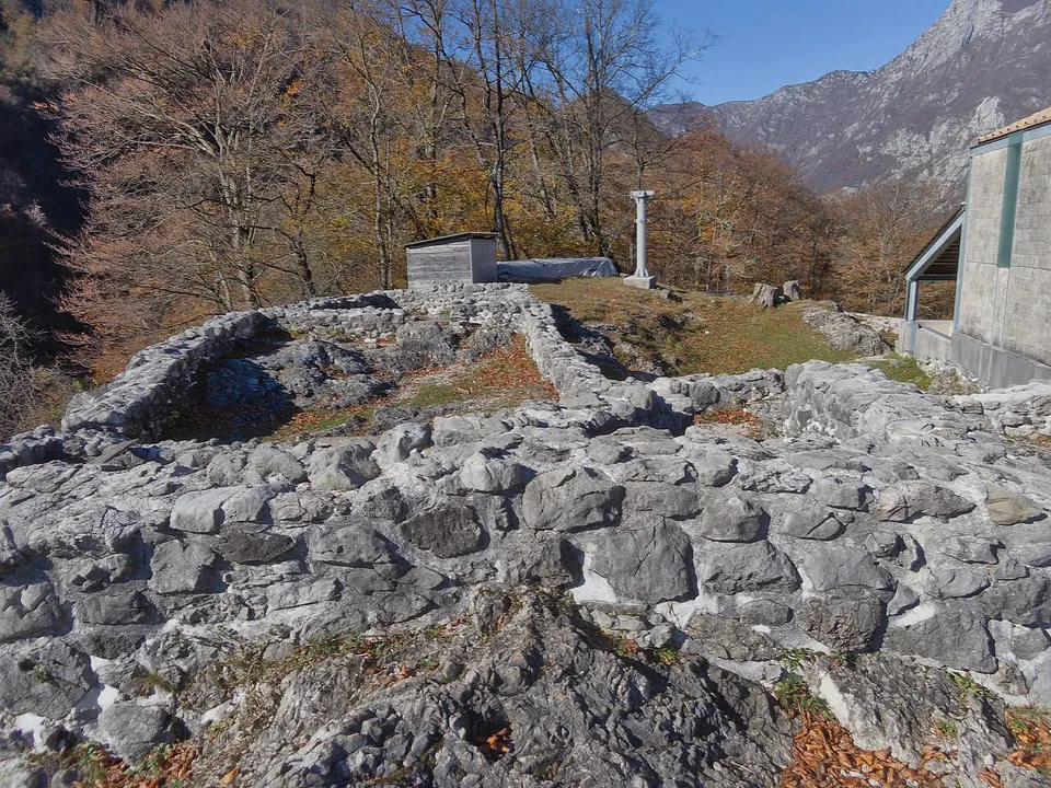Close view of rough grey limestone walling set on bare rock, with a white stone post and a small grey building behind and autumn trees and a mountain face beyond.