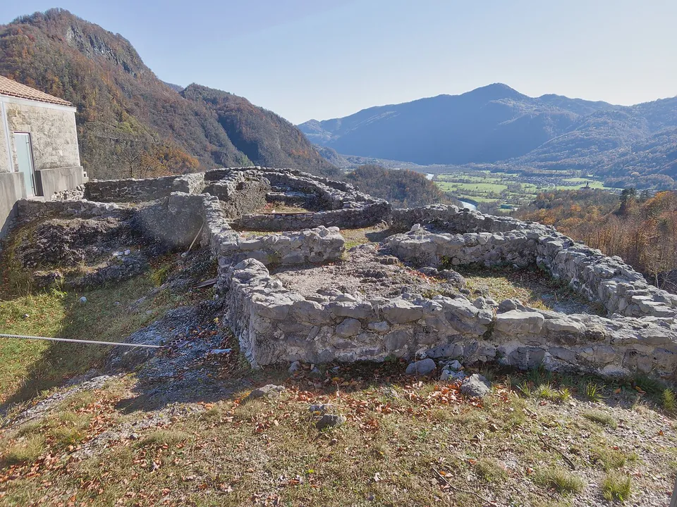 Low rebuilt stone walls of several rooms on the edge of a hilltop, with a small stone building to one side and a wide green valley and mountains beyond.