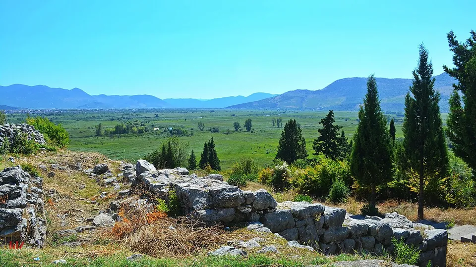 Tumbled grey stone wall blocks along a dry hillside in the foreground, looking out over a wide green river plain with cypress trees and mountains on the horizon.