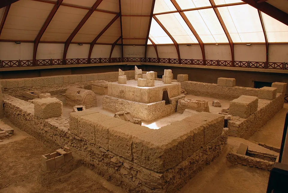 A square stone structure with thick walls and internal compartments, excavated below floor level and covered by a modern tent roof of pale fabric on steel arches, with a visitors' walkway running round it.