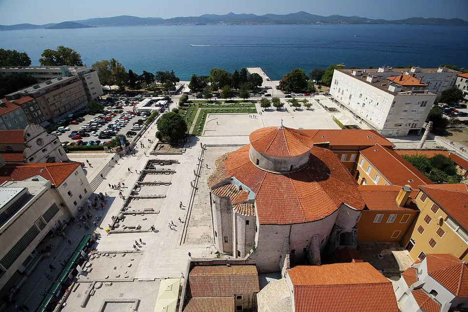 Looking down from a height on a round church with a domed red roof beside a large open paved square scattered with low stone remains, with a car park and the sea beyond.